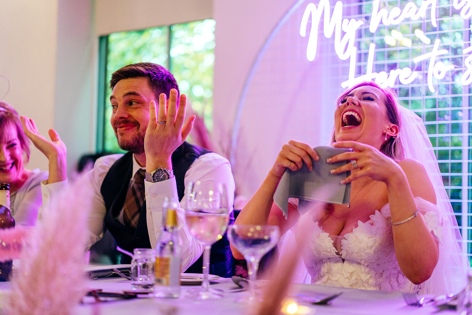 Bride throws head back laughing during speeches whilst groom grimaces 