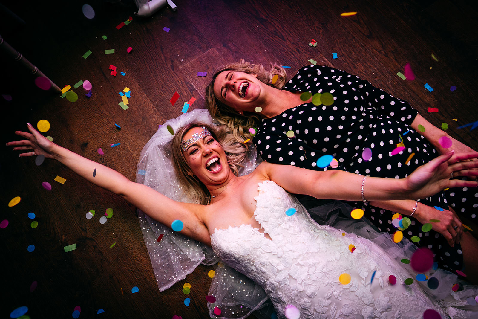 Bride laying on floor with one of her best friends throwing confetti in the air 