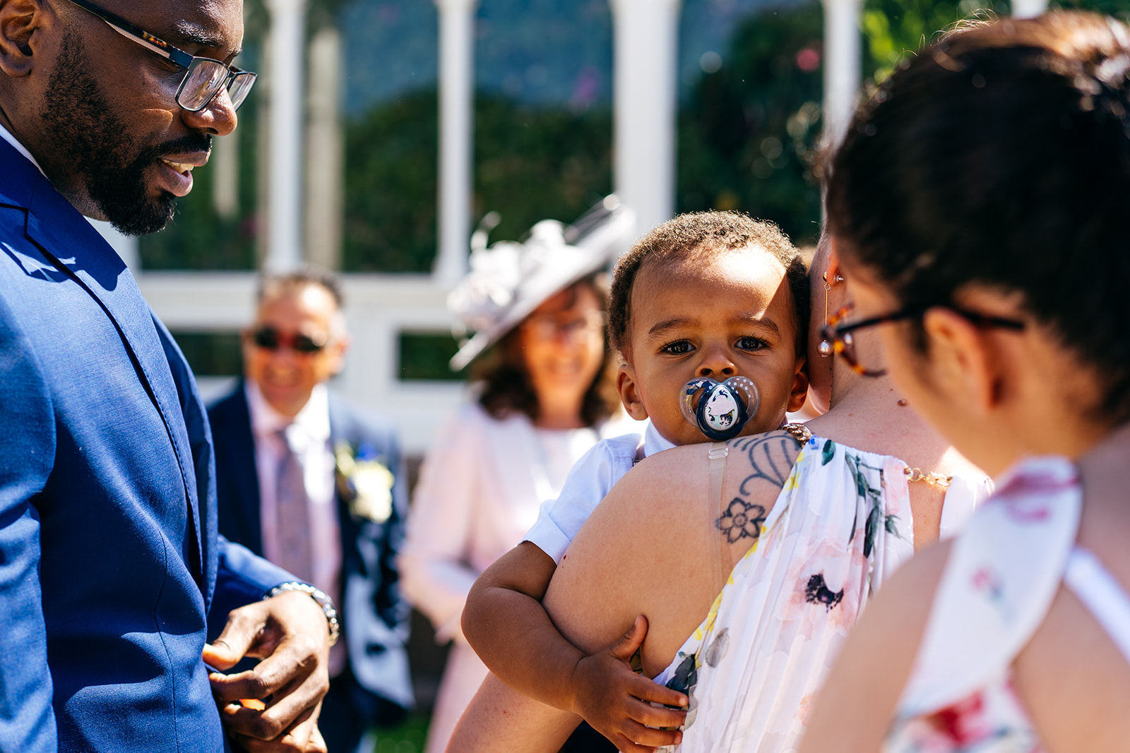 Wedding guests arrive at Sefton Park Palm House. Baby with dummy looks straight down my lens over his mother's shoulder. 