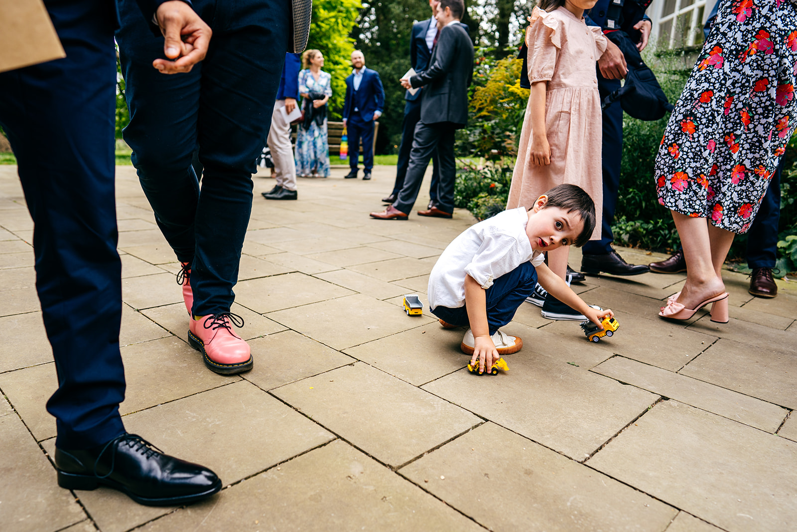 Little boy plays with his car amongst legs of adult guests waiting to enter wedding at Orleans House in London. 