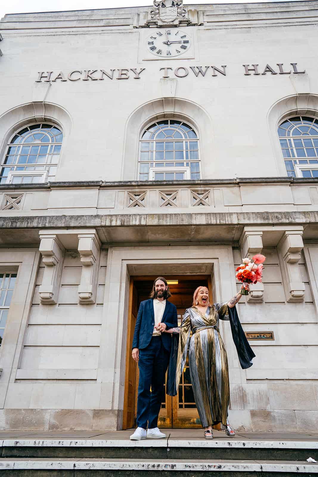 Emma and her eight bridesmaids (and two babies) in shades of red, orange and hot pink outside the Prince Albert in Camden, London. They're holding bouquets of orange and yellow flowers with lots of foliage and bright ribbons. Credit Jordanna Marston.